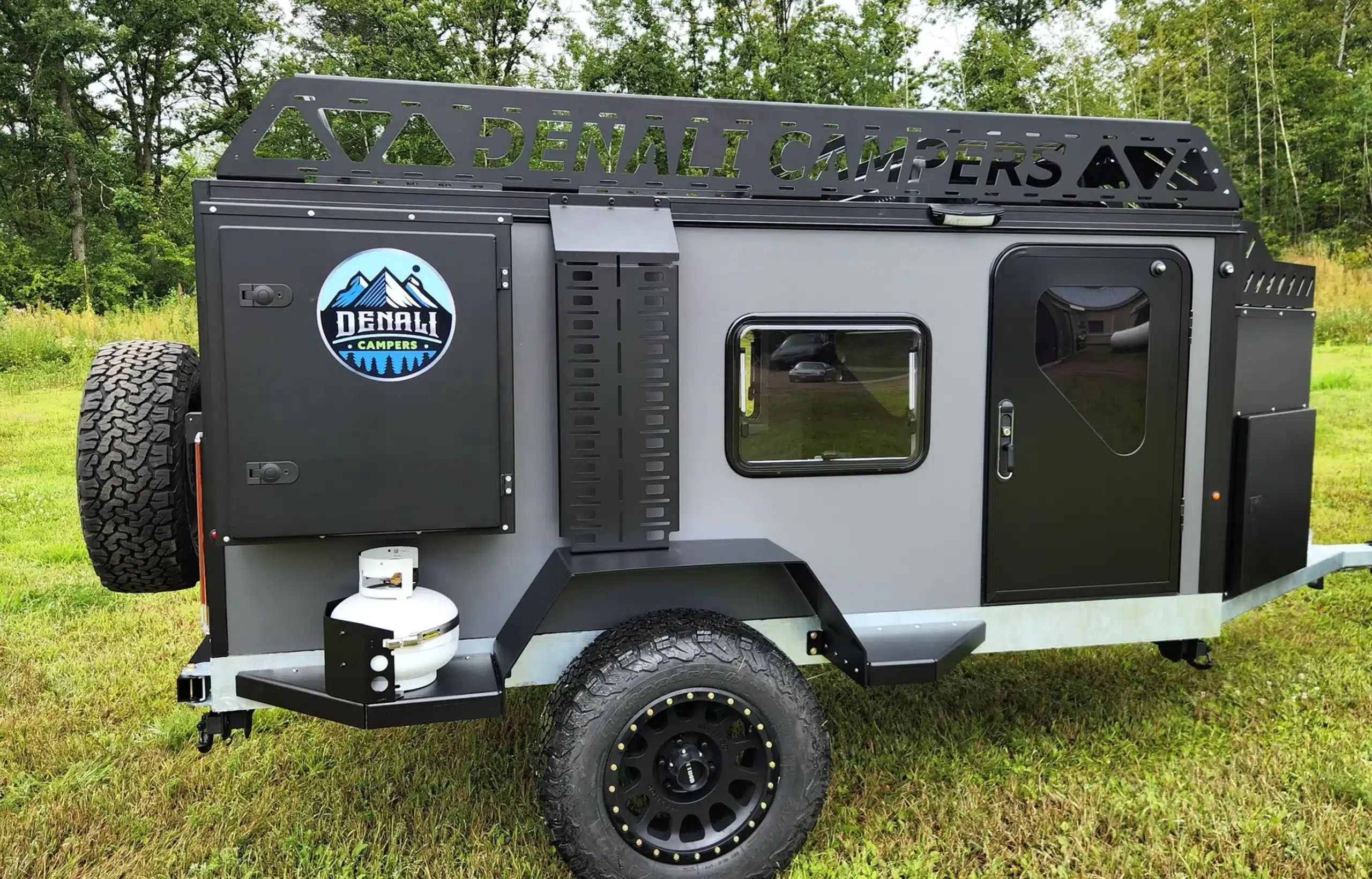A compact Denali Camper XRT-10 trailer with rugged tires, a black and gray exterior, a mounted spare tire, and a front storage box is parked on grass with trees in the background.