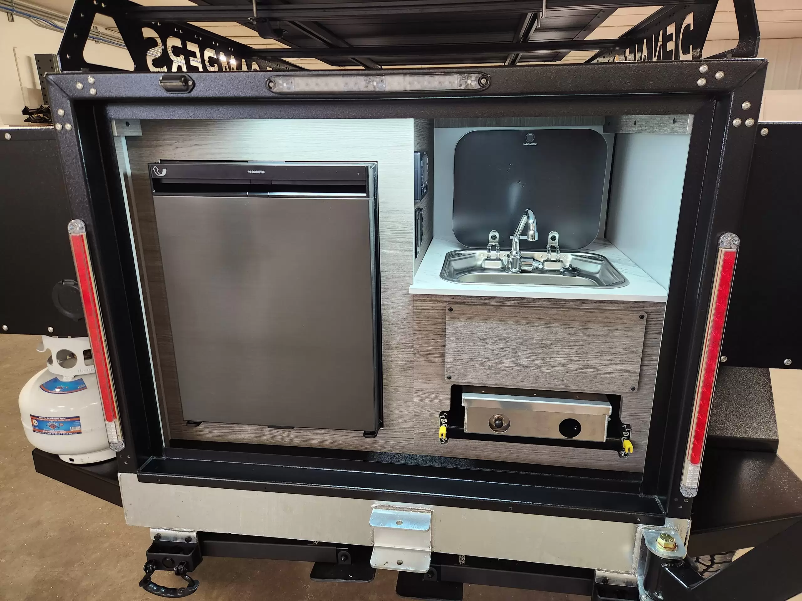 A compact outdoor kitchen by Denali Campers is built into the back of a truck or trailer, making travel effortless. With a mini fridge, sink, stove, storage drawers, and side-mounted propane tank, it's easy to set up for on-the-go adventures.
