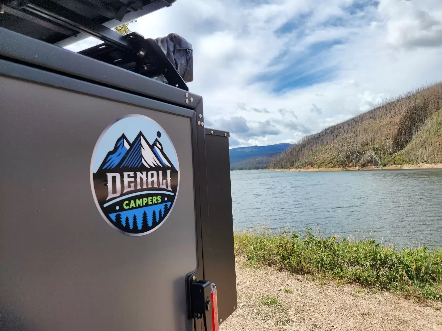 A Denali Campers logo on a vehicle is in the foreground, with a scenic lake, mountains, and a partly cloudy sky in the background—perfect for those seeking off grid adventures. Green grass and gravel line the water’s edge.