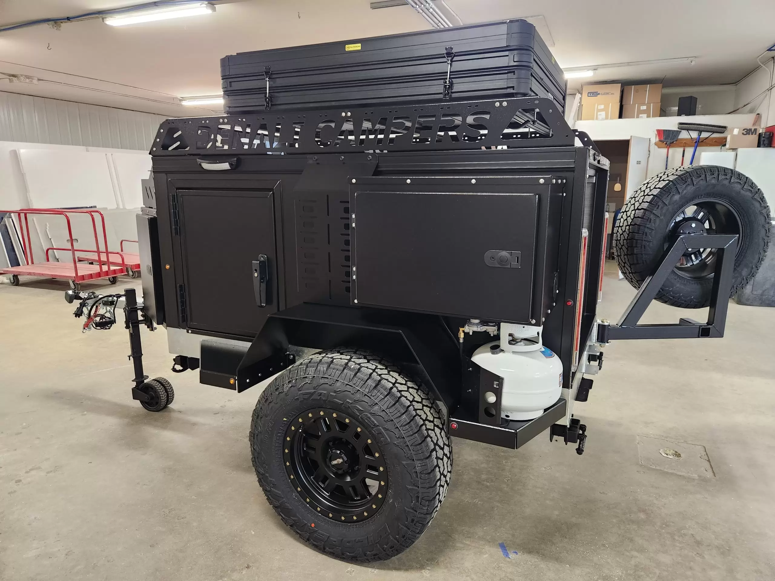 A black Denali Camper off-road trailer with rugged tires, storage compartments, a mounted spare tire, propane tank, and roof cargo box is displayed indoors in a garage or workshop setting.