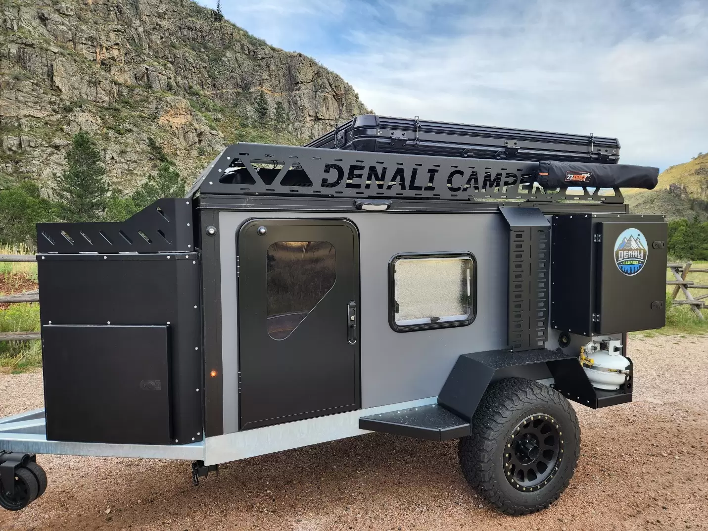 A rugged black and silver Denali Campers trailer is parked outdoors near rocky cliffs and greenery, featuring off-road tires, a roof rack, a rooftop tent, and a side window—perfect for year-round adventure.