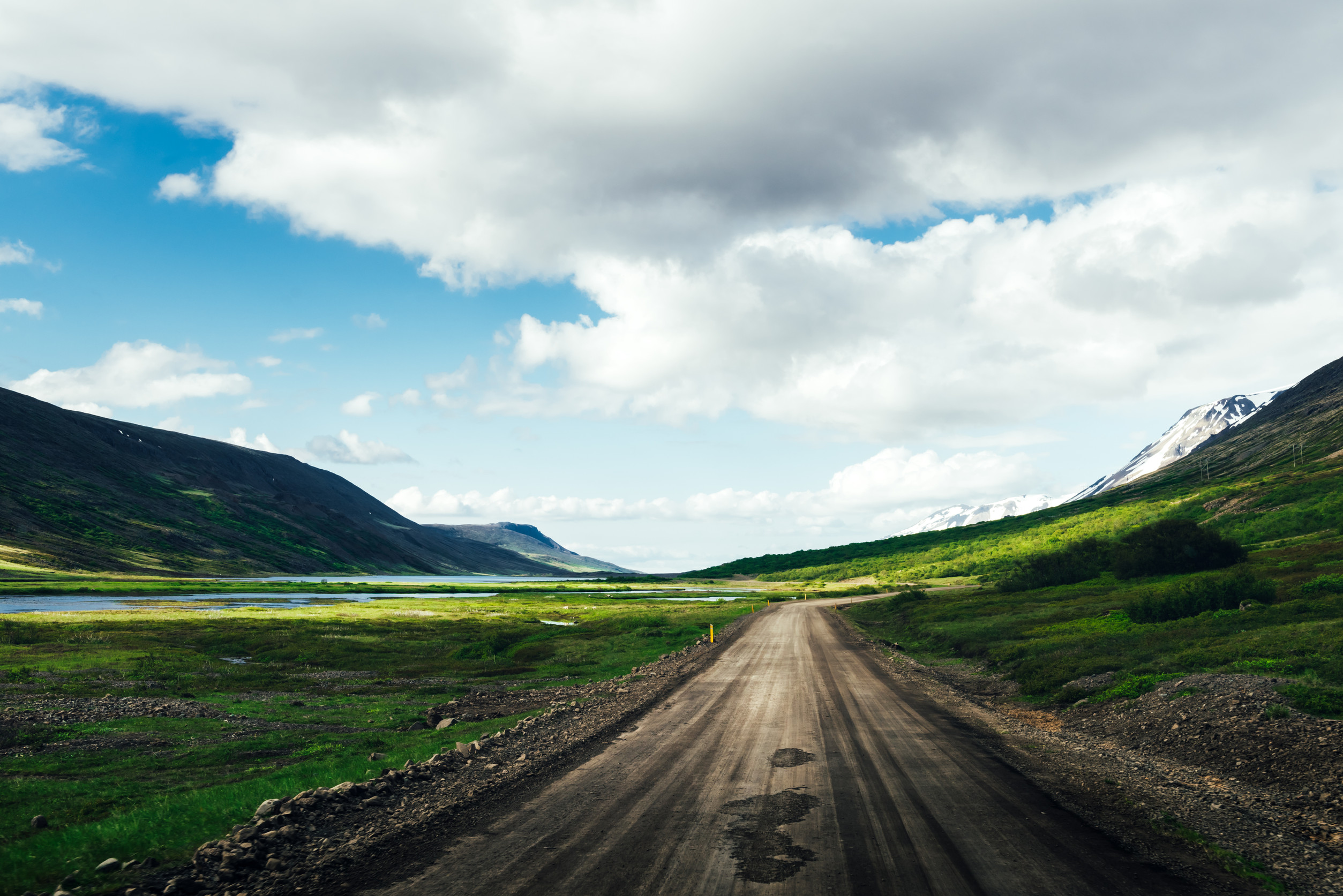 A dirt road winds through a lush green valley with mountains on both sides, a river to the left, and a partly cloudy sky overhead—an ideal setting for off-road camper trailers and endless adventure.